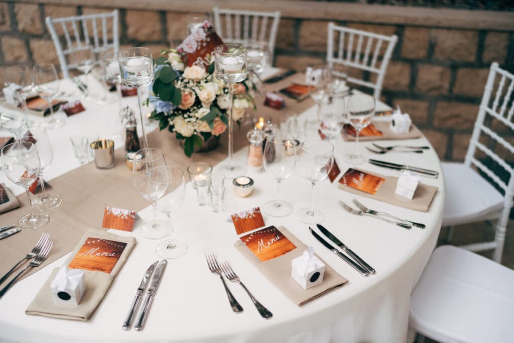 Round table with a white tablecloth with a brown runner on the table. Vienna nested in brown napkins, a bouquet of flowers is on the table. White Chiavari chairs with pillows.