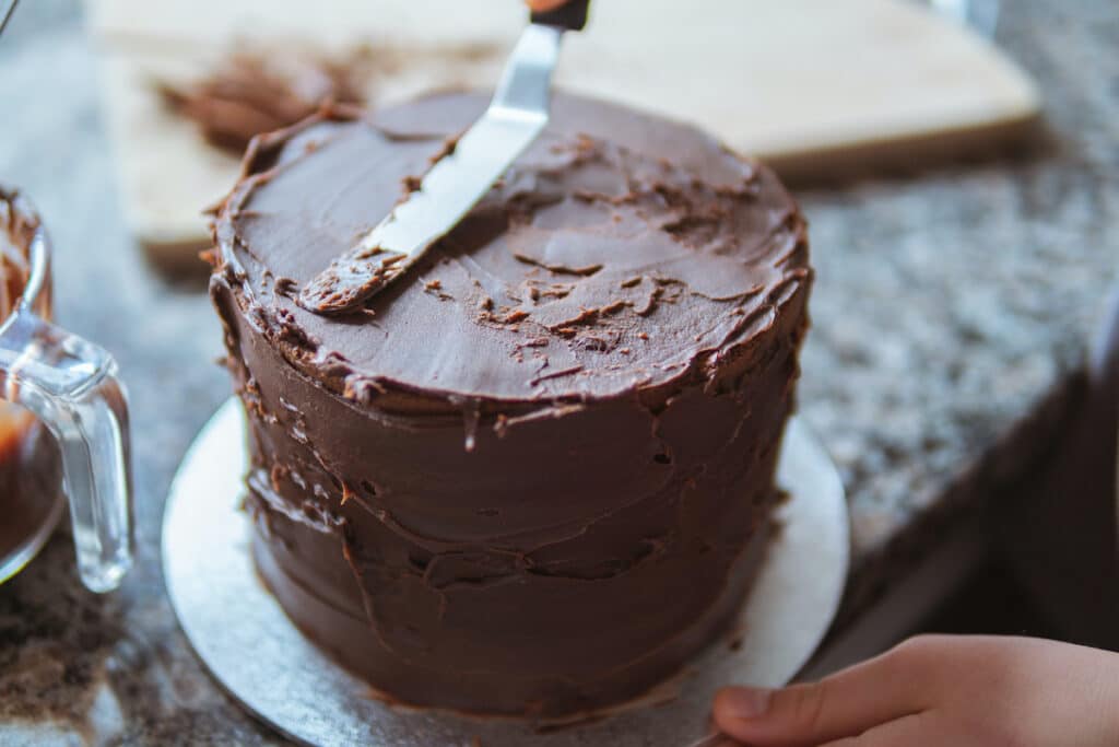 A close-up photograph of a girl decorating a cake with chocolate ganache in a domestic kitchen