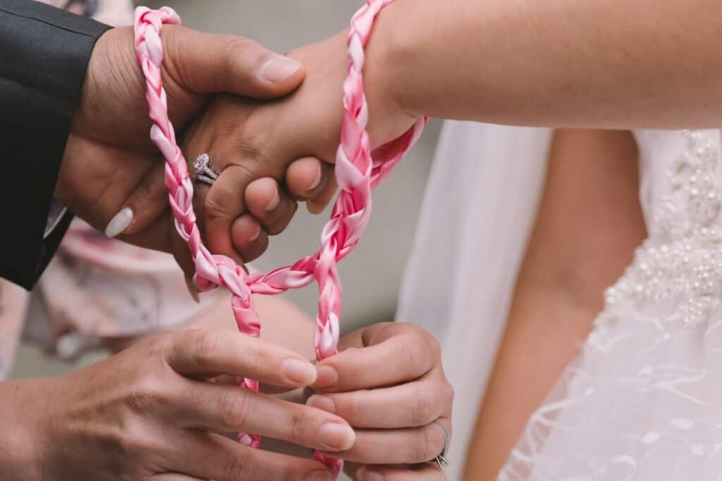 Bride and groom holding each others left hand with red and white handfasting cord wrapped around their wrists 