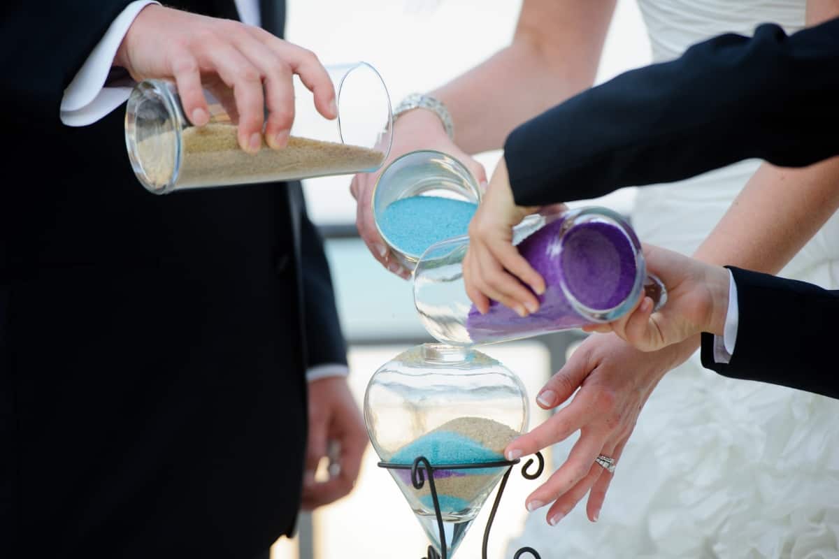 Three hands pour purple, blue and gold sand into glass bowl during sand ceremony